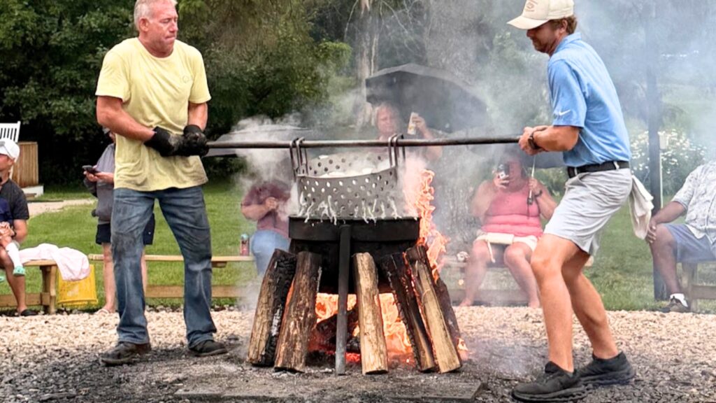 Two people lift a large metal basket over an outdoor wood fire in scenic Door County, while several people sit and watch in the background.