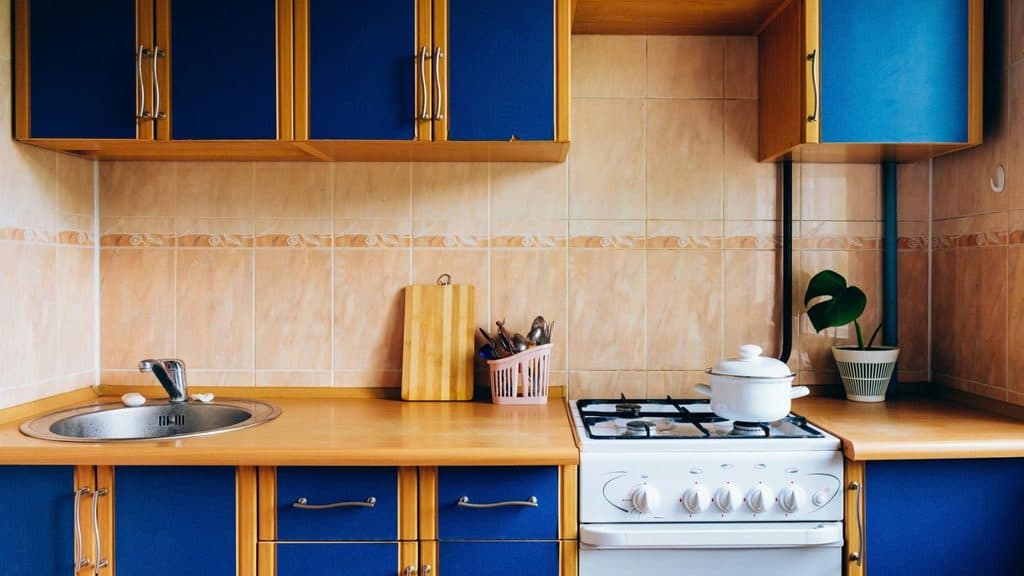 Modern kitchen with blue and wood cabinets, a gas stove with a pot, classic kitchen gear Americans remember, a sink, a chopping board, utensils in a basket, and a potted plant on the countertop.
