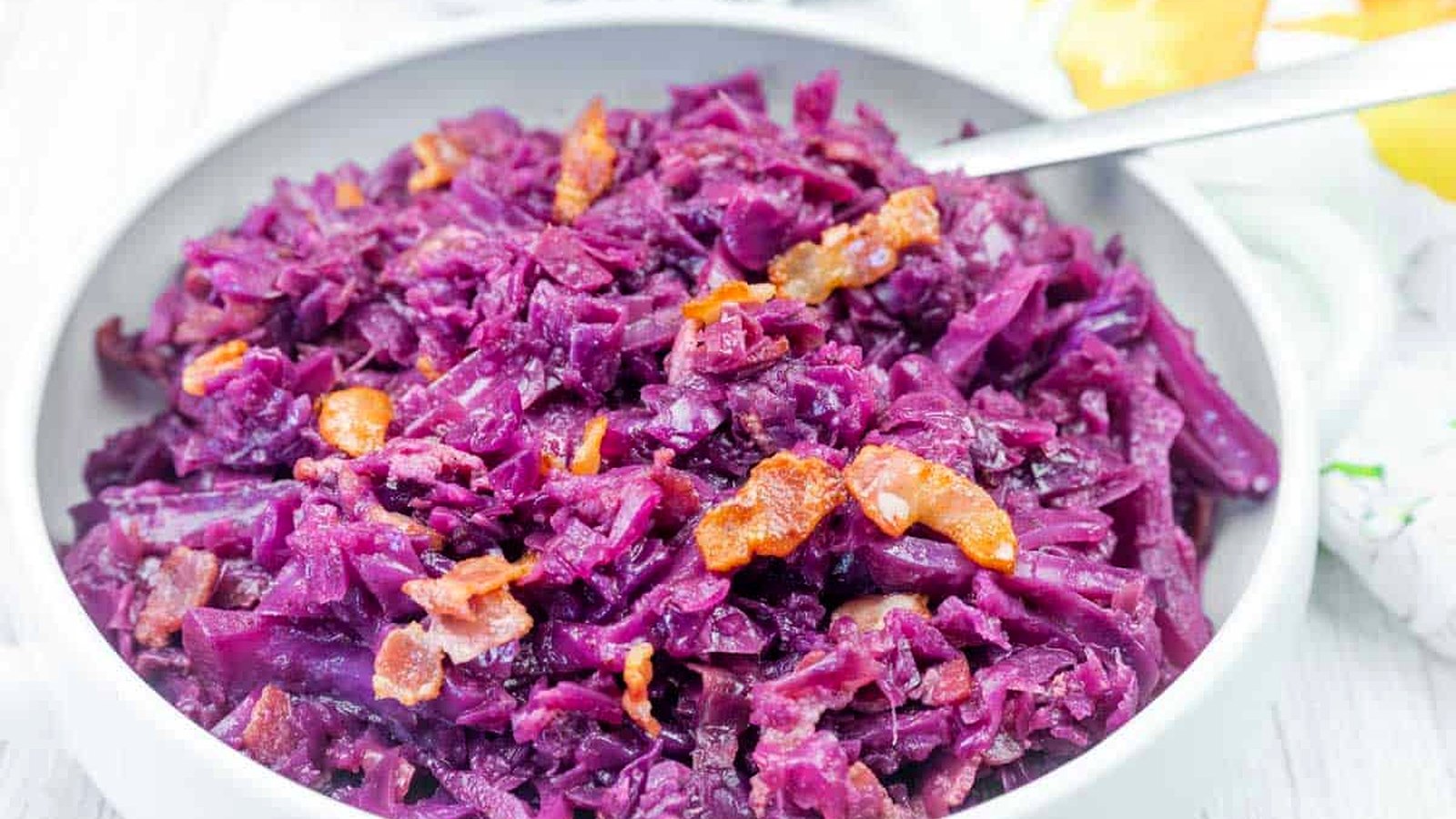 A bowl of cooked red cabbage mixed with small pieces of bacon, served with a spoon.