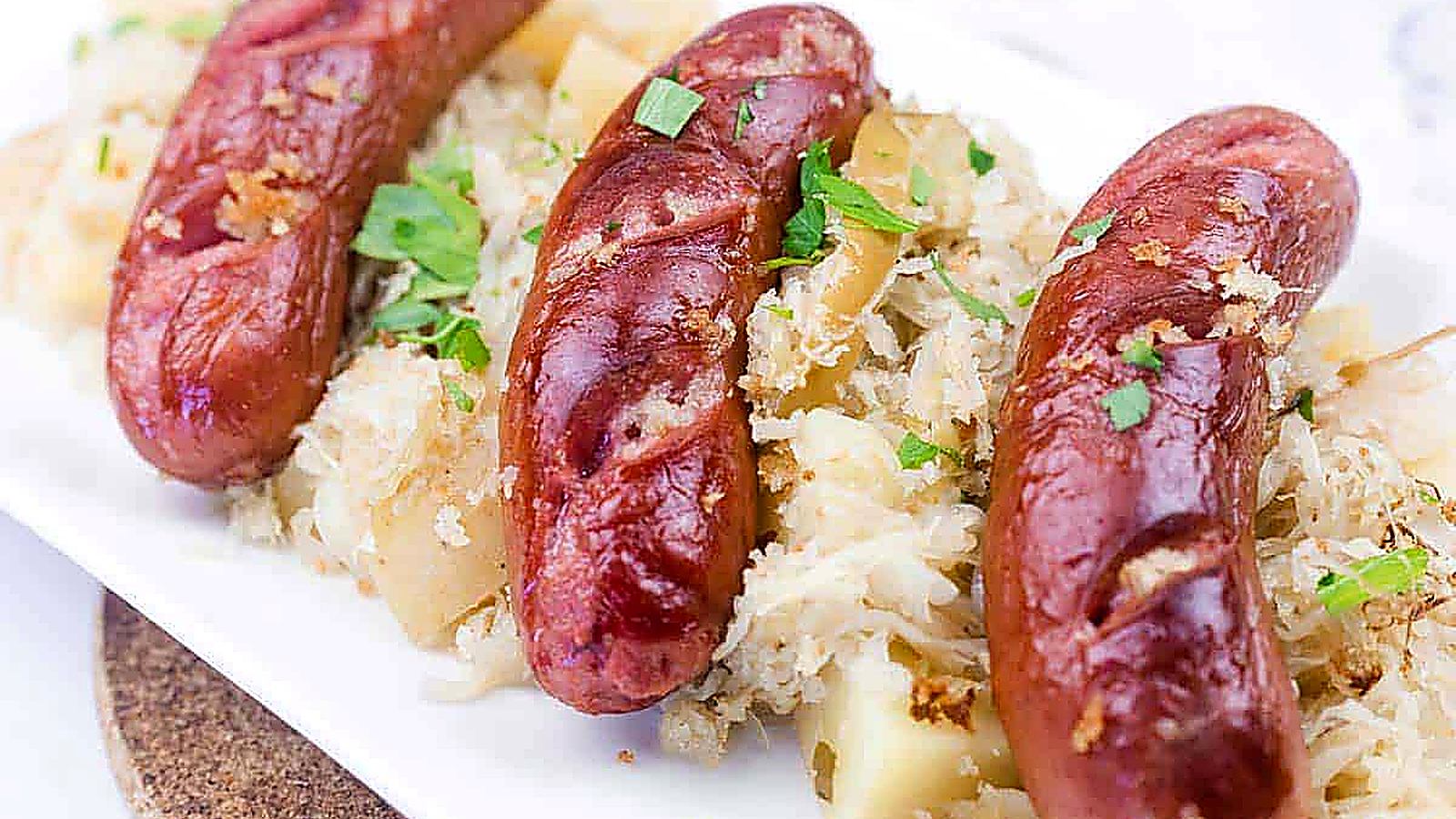 Three grilled sausages served on a bed of sauerkraut and diced potatoes, garnished with chopped parsley on a white plate.