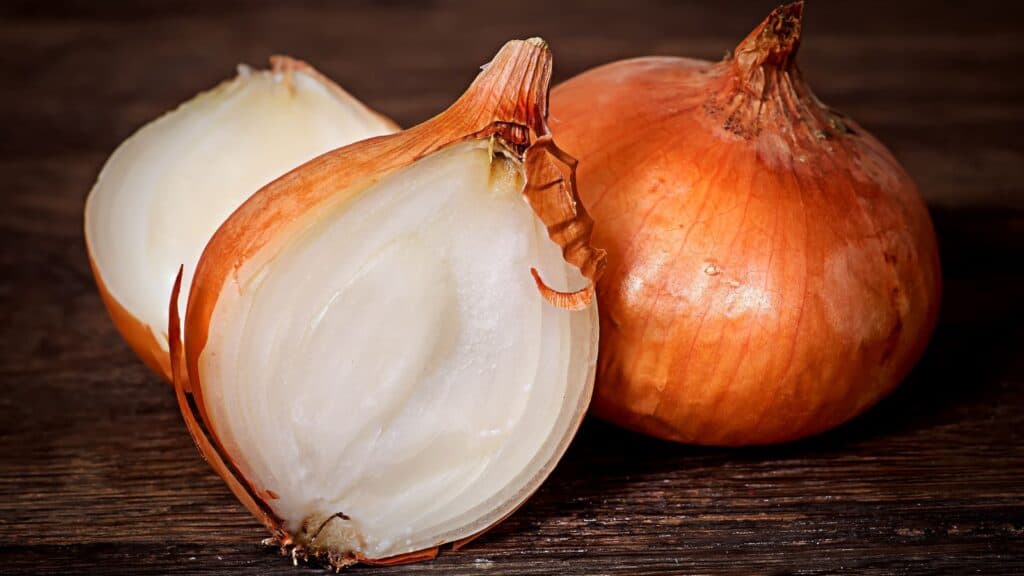 A whole brown onion and a halved onion are placed on a wooden surface, showing the white interior and papery brown skin&mdash;an everyday ingredient that, in recent years, has been among foods linked to major recalls.