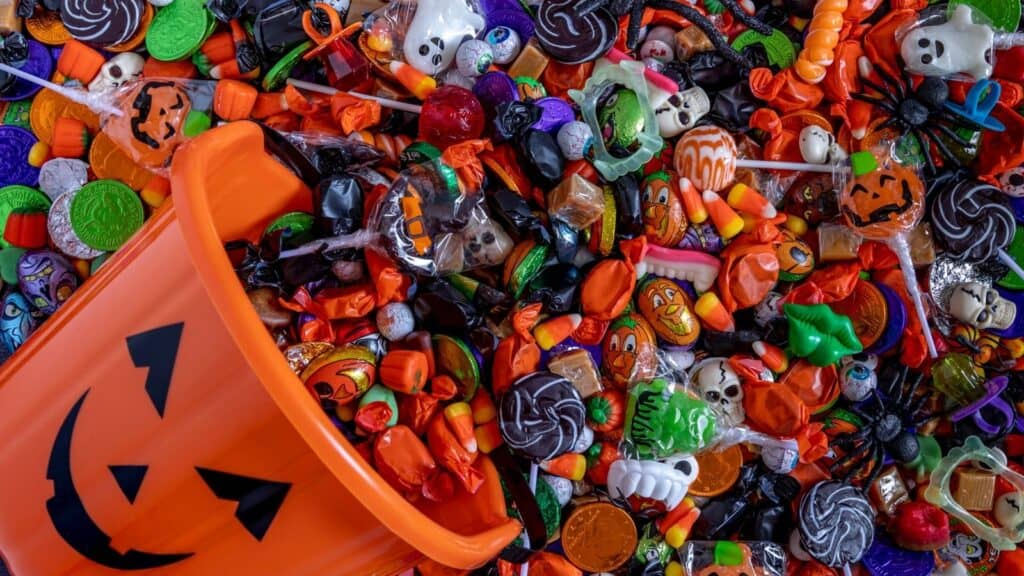 Orange pumpkin bucket spilled with assorted Halloween candies, chocolates, and small toys in festive Halloween-themed wrappers and shapes.