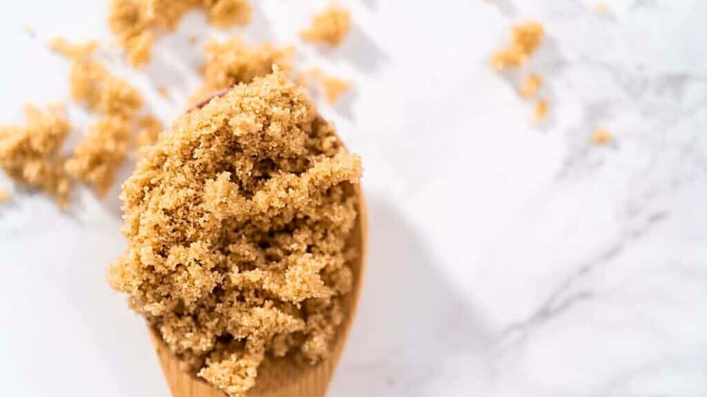 A wooden spoon holds a heaping scoop of light brown sugar, with small clumps of sugar scattered on a white marble surface.