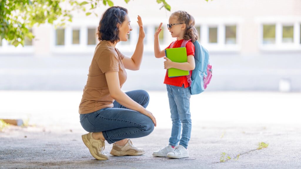 An adult crouches to give a high-five to a child wearing a backpack and glasses, who is holding notebooks and standing outside—capturing the excitement of back to school budgets in action.