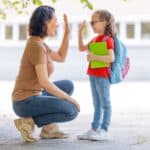 An adult crouches to give a high-five to a child wearing a backpack and glasses, who is holding notebooks and standing outside&mdash;capturing the excitement of back to school budgets in action.