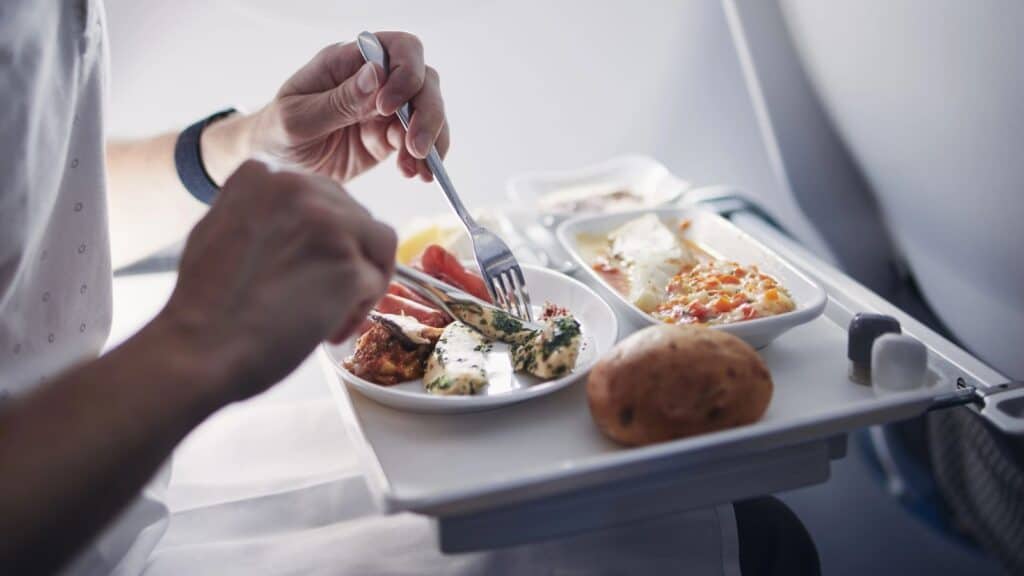 A person eating a meal on an airplane, with food trays containing vegetables, a bread roll, and various dishes on a fold-down tray table.