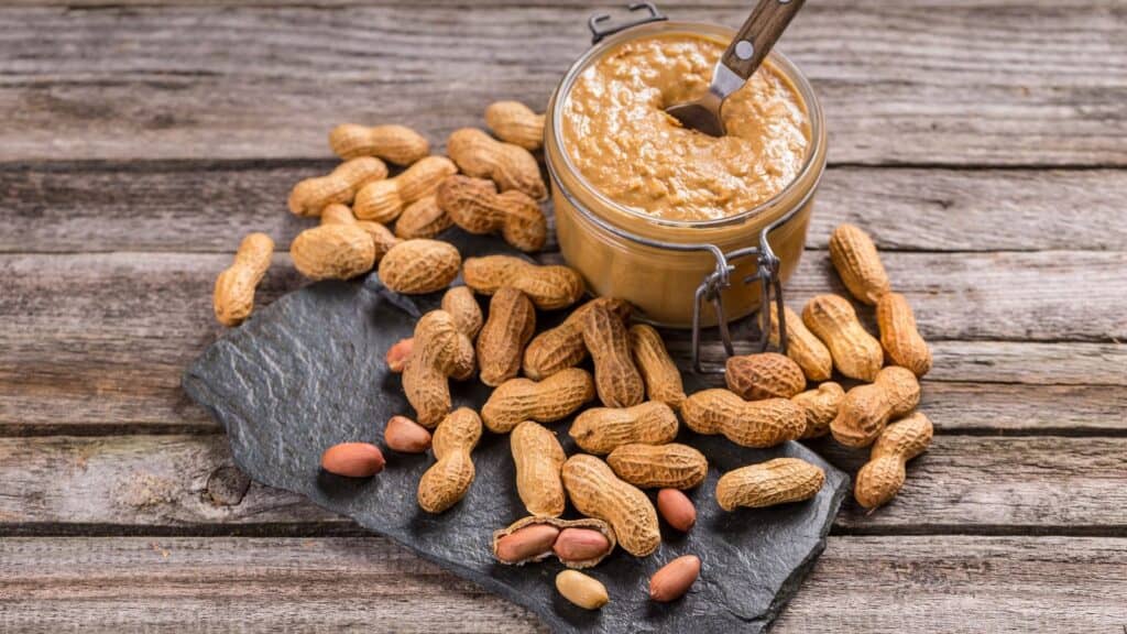 A jar of peanut butter with a spoon surrounded by whole peanuts in shells on a slate and wooden surface.