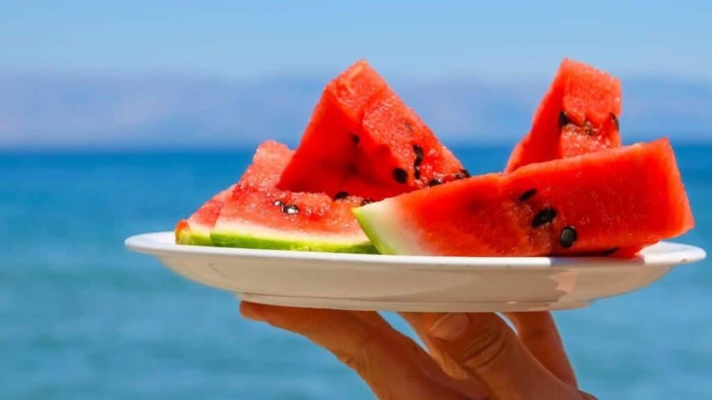 A hand holds a white plate with several slices of watermelon&mdash;one of the foods risky in summer heat&mdash;in front of a blue sea and sky background.