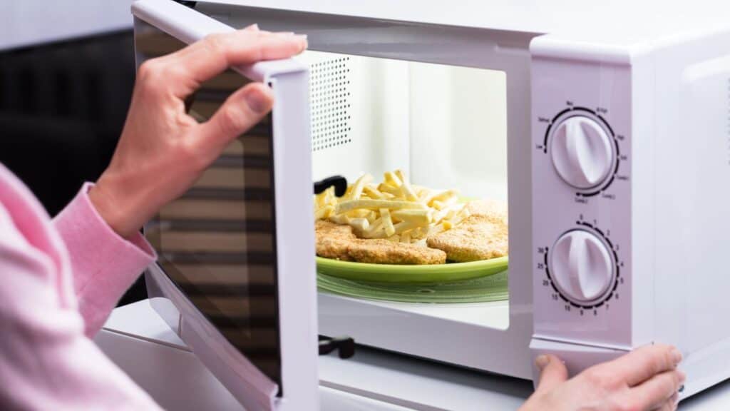 A person places a plate with breaded food and fries into a microwave, preparing to heat them up using kitchen gear Americans remember.