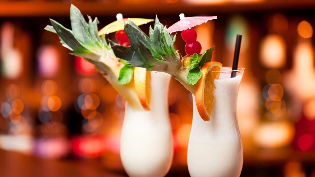 Two pina colada cocktails in curved glasses, garnished with pineapple, orange slices, cherries, mint, and small umbrellas, set against a blurred bar background.