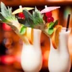 Two pina colada cocktails in curved glasses, garnished with pineapple, orange slices, cherries, mint, and small umbrellas, set against a blurred bar background.