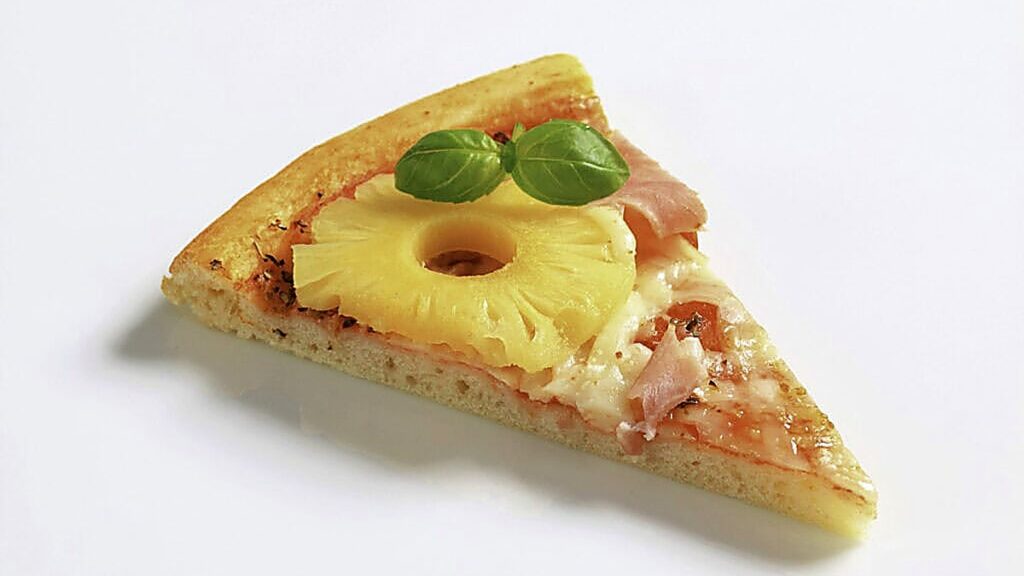 A slice of pizza topped with ham, pineapple, melted cheese, and two fresh basil leaves on a plain white background.