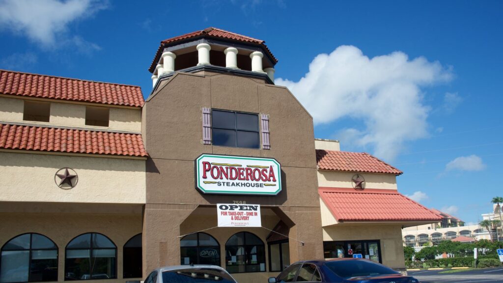 Ponderosa Steakhouse exterior with a large sign, arched entrance, red tile roof, and several parked cars under a blue sky with clouds—one of the classic salad bar chains known for inviting families to dine in comfort.