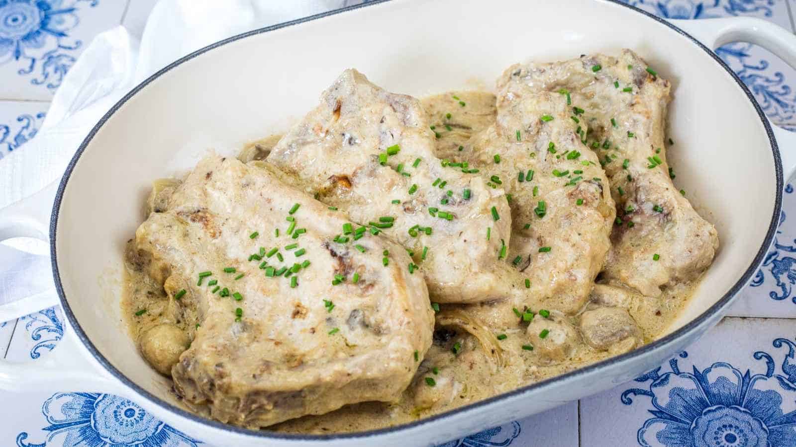 Four pork chops in a creamy white sauce, garnished with chopped chives, served in a white oval dish on a blue and white tiled surface.