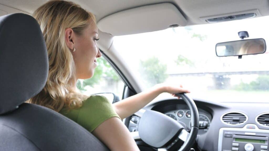 A woman with blonde hair sits in the driver&rsquo;s seat of a car&mdash;perhaps contemplating how unexpected rental car fees catch travelers unaware&mdash;holding the steering wheel and looking forward through the windshield.