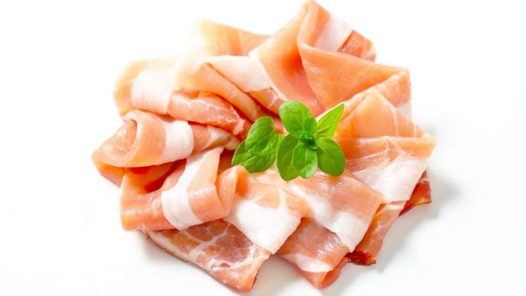 Thin slices of prosciutto, one of the renowned foods with protected names in Europe, are arranged in a pile on a white surface, garnished with a small sprig of green basil.