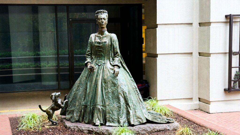Bronze statue of a woman in a long dress holding flowers, with a small dog beside her, situated on a stone base outside a building&mdash;a charming landmark that highlights the city&rsquo;s unique culture and contributes to Charlotte tourism spending.