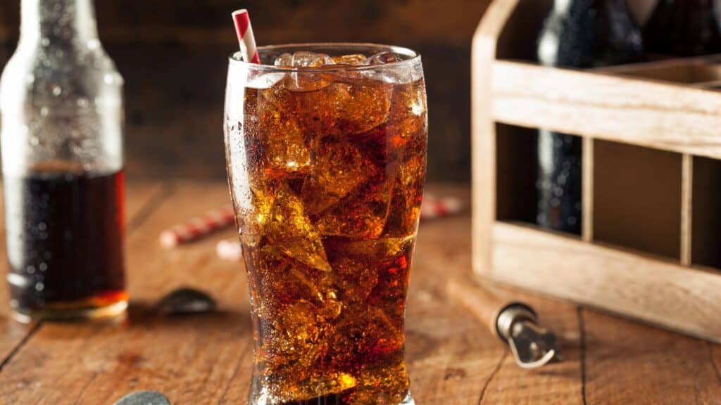 A glass of cola with ice and a straw sits on a wooden table, with a bottle, bottle opener, and soda carrier in the background&mdash;a nostalgic nod to sodas that had their moment.