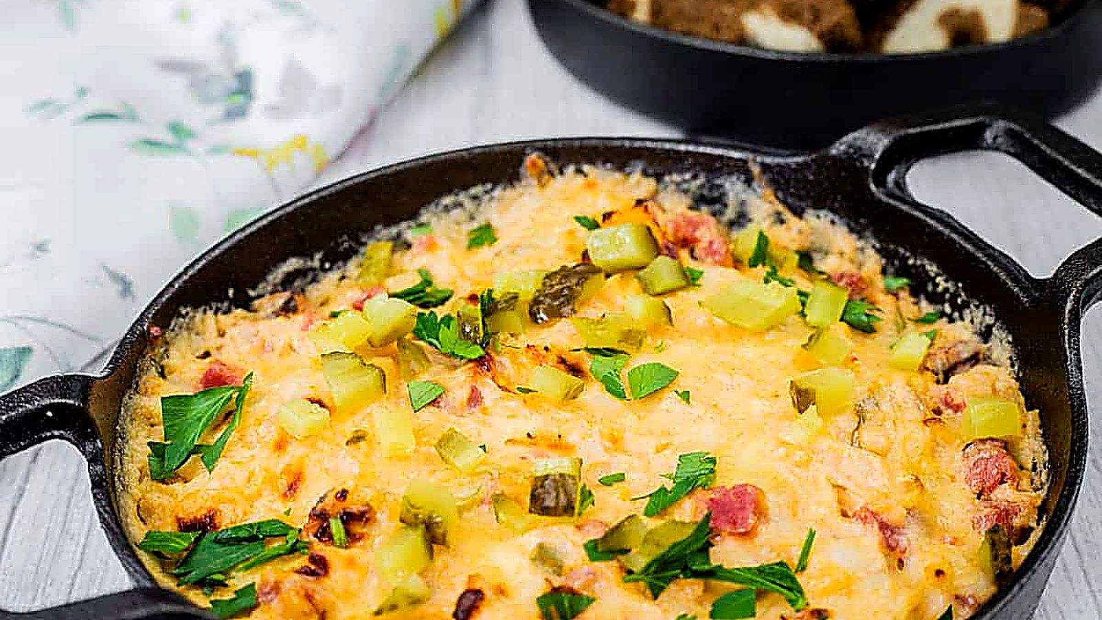 Cheesy, golden-brown Hot Reuben dip in a cast-iron skillet, topped with chopped pickles and fresh parsley, served with slices of marbled rye bread.