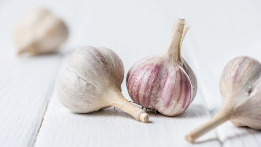 Three whole garlic bulbs rest on a white wooden surface, with two focused in the foreground and one blurred in the background—a simple reminder that not all foods you are refrigerating need to be stored cold.