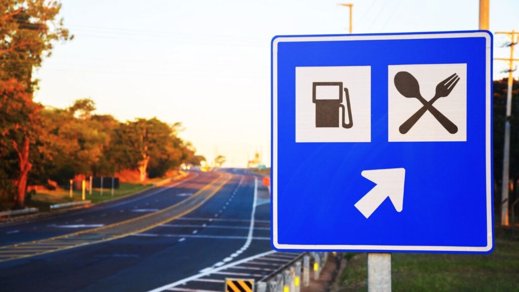 Blue road sign showing icons for fuel and food, with a white arrow pointing to the right; empty highway and trees visible in the background—a reminder that regional chains rarely cross state lines.