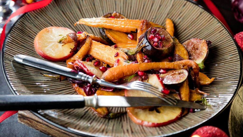 A plate of roasted carrots, figs, apple slices, and pomegranate seeds, with a knife and fork resting on the edge.