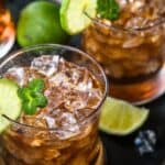 A glass filled with iced brown cocktail, garnished with a lime slice and mint, with whole and cut limes in the background.