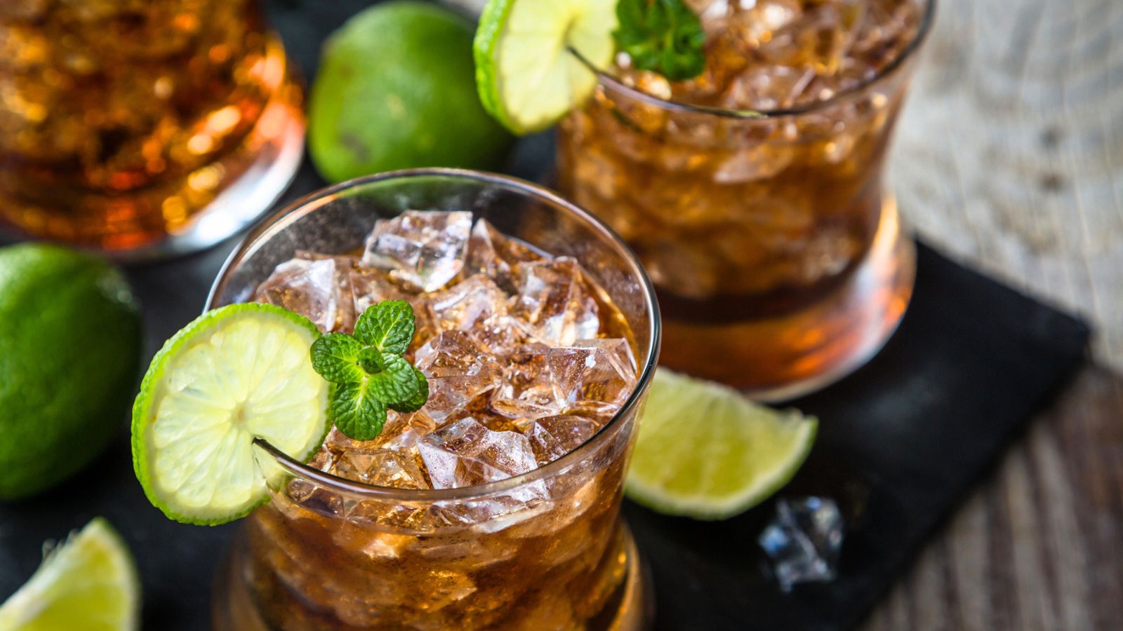 A glass filled with iced brown cocktail, garnished with a lime slice and mint, with whole and cut limes in the background.