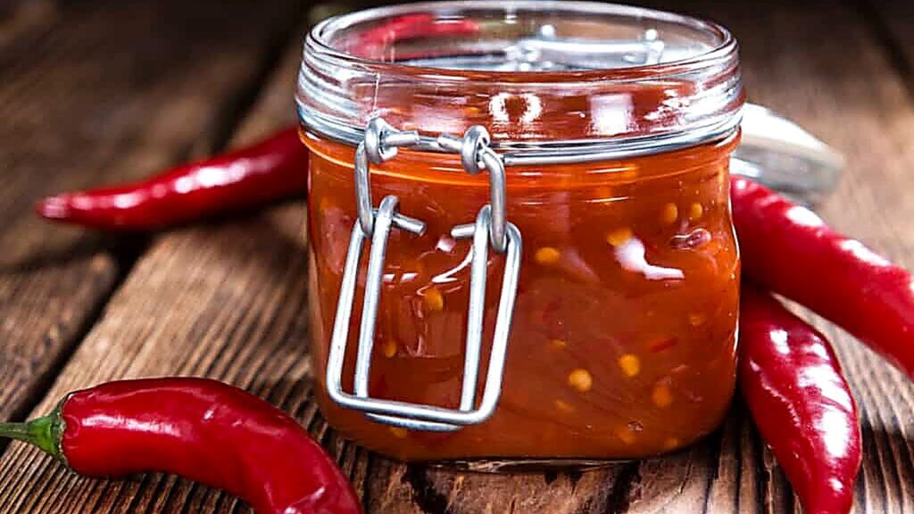 A glass jar filled with red chili sauce sits on a wooden surface, surrounded by whole red chili peppers.