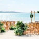 Rows of wooden chairs and green plant decorations are arranged on a sandy beach facing a wooden wedding arch, with the ocean visible in the background.
