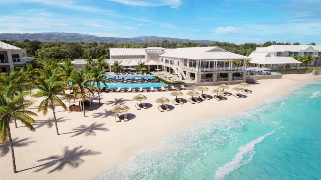 A beachfront resort with palm trees, sun loungers, straw umbrellas, a pool, and white buildings bordering a turquoise ocean under a clear sky.