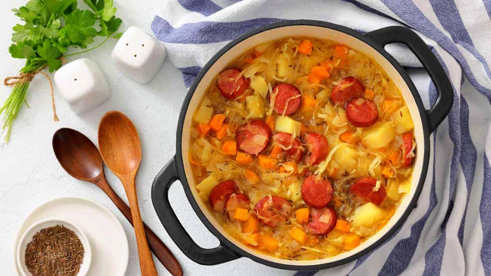 A pot of Chef Jenn&rsquo;s Sauerkraut Soup with Kielbasa sits on a striped towel next to wooden spoons, bowls, fresh herbs, and salt and pepper shakers.
