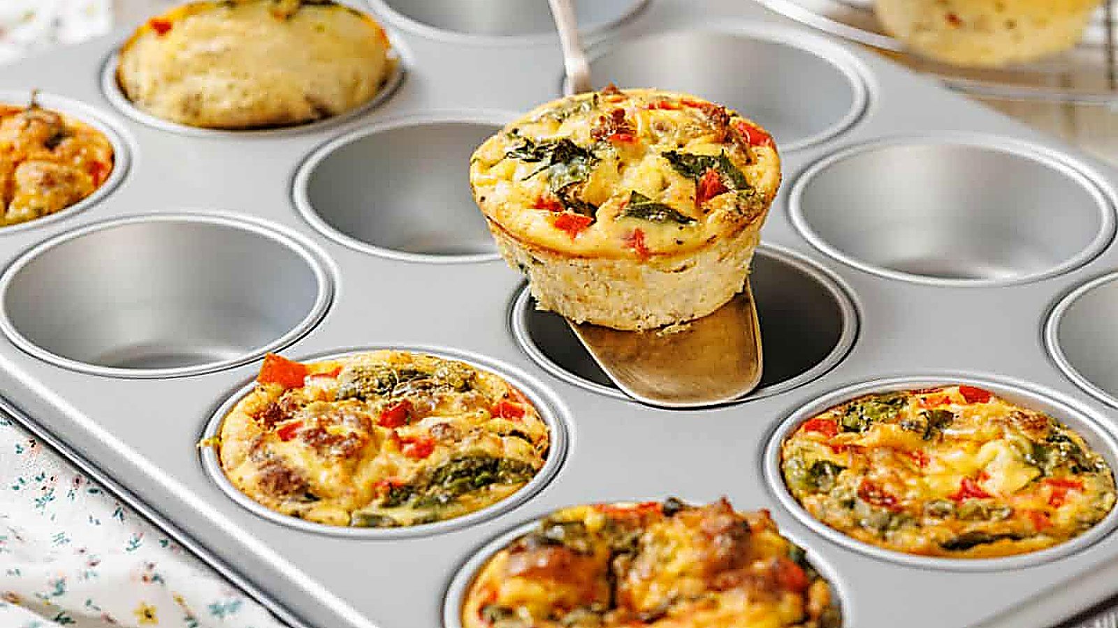 A metal muffin tin holds several breakfast baked egg muffins with vegetables; one is being lifted out with a spatula.