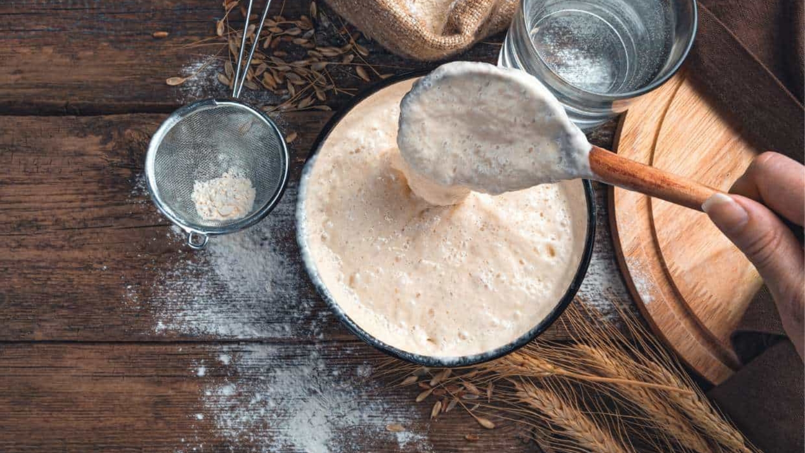 A hand stirs bubbly sourdough starter in a bowl on a wooden table, with flour, a strainer, wheat stalks, and a glass of water nearby.