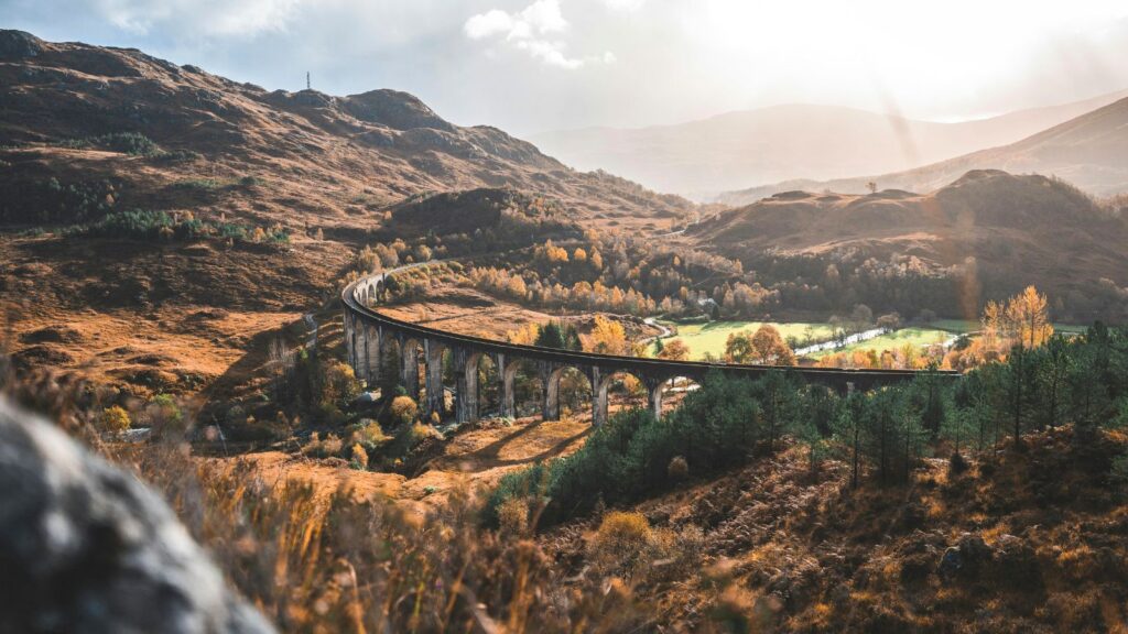 A curved stone viaduct crosses a sunlit valley with hills, autumn trees, and grassy fields in the background&mdash;a scene perfect for those seeking inspiration on their Routescape luxury travel adventures.