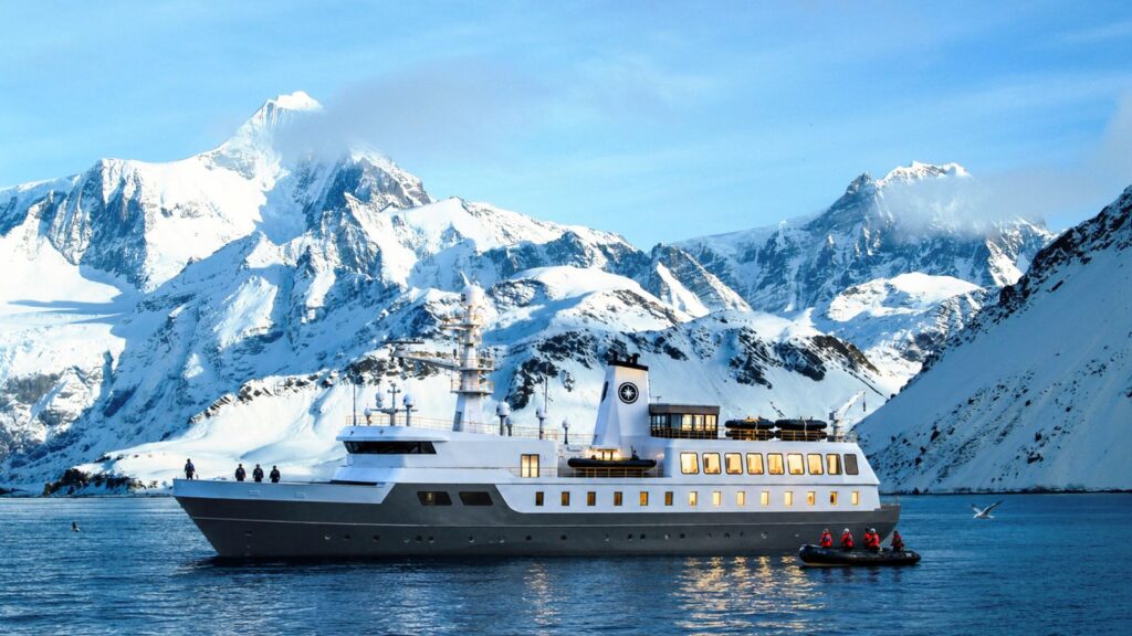 A large white yacht is anchored on calm water near snow-covered mountains, with people in a small boat nearby.