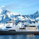 A large white yacht is anchored on calm water near snow-covered mountains, with people in a small boat nearby.