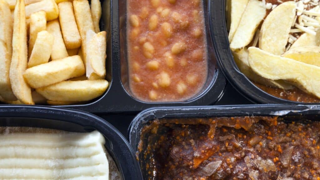 Close-up of a frozen dinner tray, shaped in the classic American freezer style, featuring baked beans, French fries, mashed potatoes, potato wedges with cheese, and a portion of minced meat in sauce.