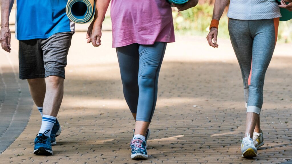 Three adults in athletic wear walk on a paved path outdoors, each carrying a rolled-up yoga mat. Only their legs and torsos are visible.