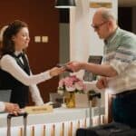 Man hands a phone to a receptionist at a hotel front desk, while another woman stands nearby with luggage and a reception sign is visible, highlighting typical US hotel fee facts guests may encounter during check-in.