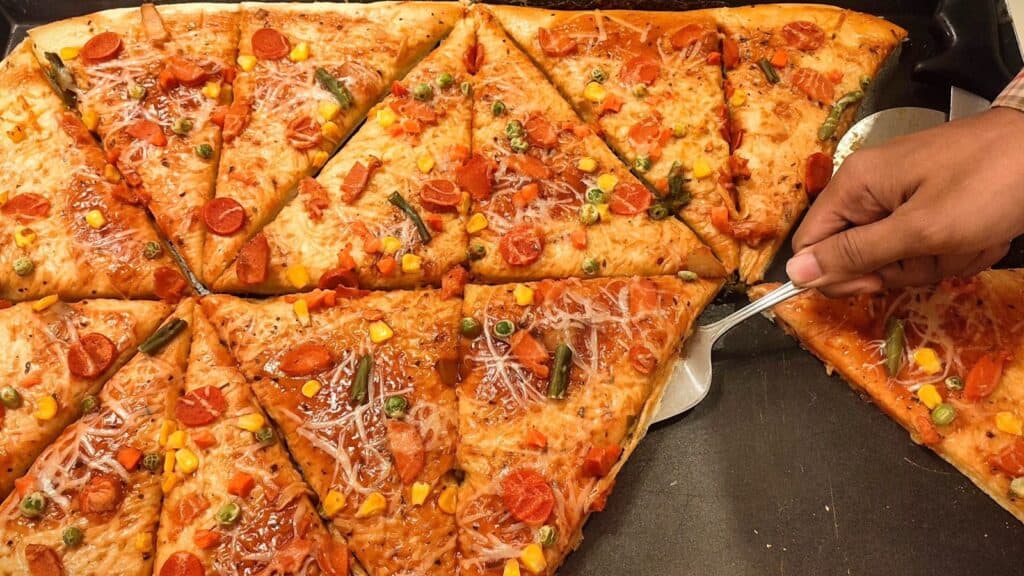 A hand uses a metal spatula to lift a slice of pizza topped with pepperoni, cheese, green beans, peas, and corn from a large tray of cut pizza slices.