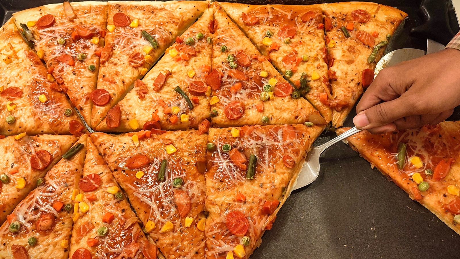 A hand uses a metal spatula to lift a slice of pizza topped with pepperoni, cheese, green beans, peas, and corn from a large tray of cut pizza slices.