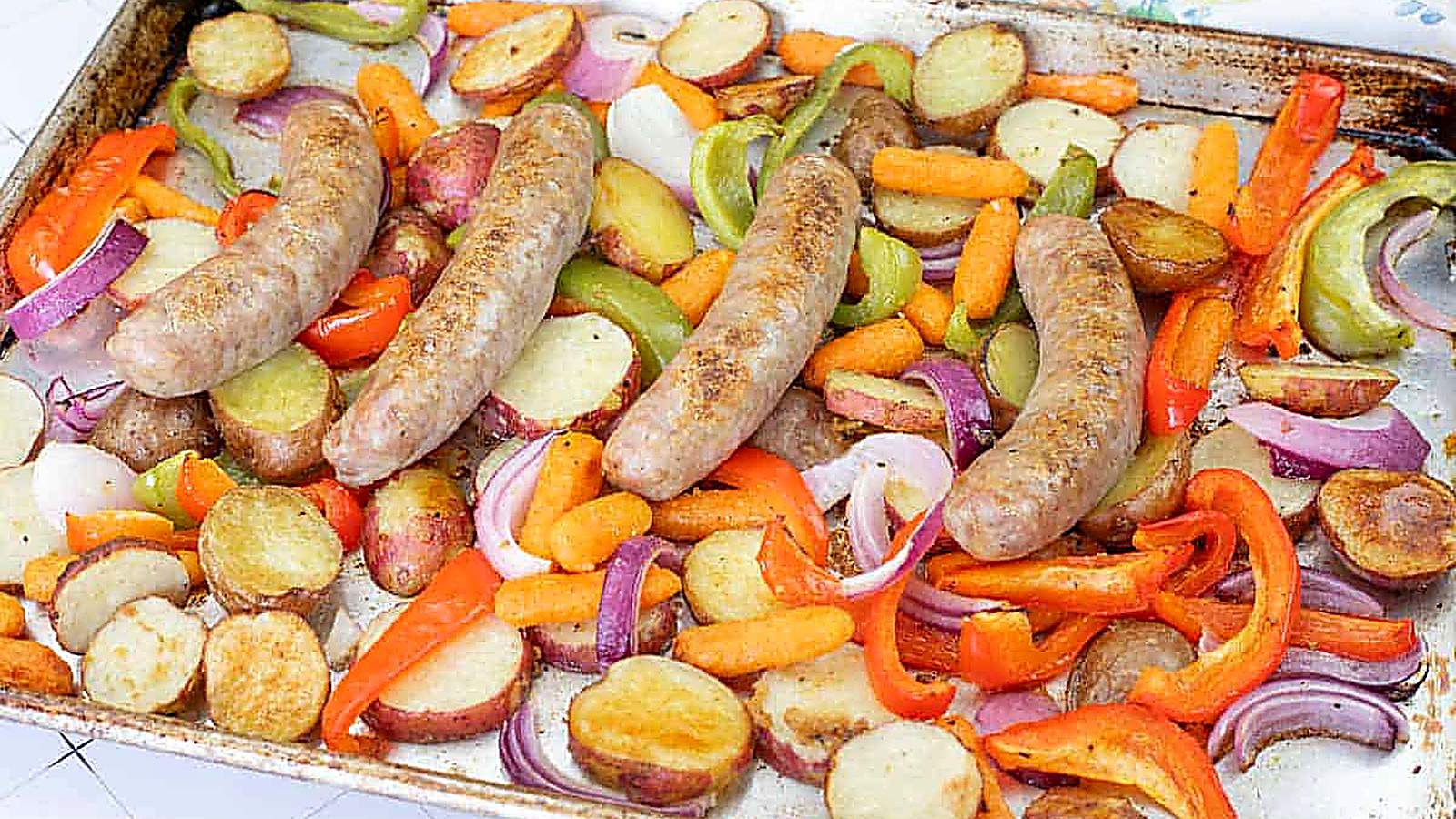 A sheet pan filled with cooked sausages, sliced potatoes, carrots, red and green bell peppers, and red onions.