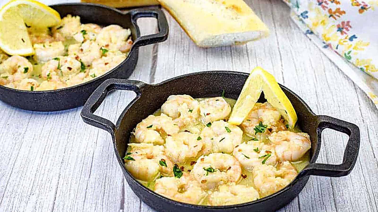 Two cast iron skillets contain shrimp scampi garnished with lemon wedges.