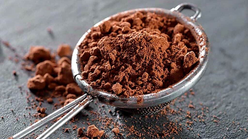 A metal sieve filled with cocoa powder sits on a dark surface, with some cocoa powder scattered around it.