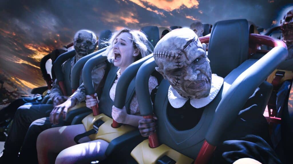 People dressed as zombies and a screaming woman ride a roller coaster at sunset during Six Flags Halloween, surrounded by dramatic clouds in the background.