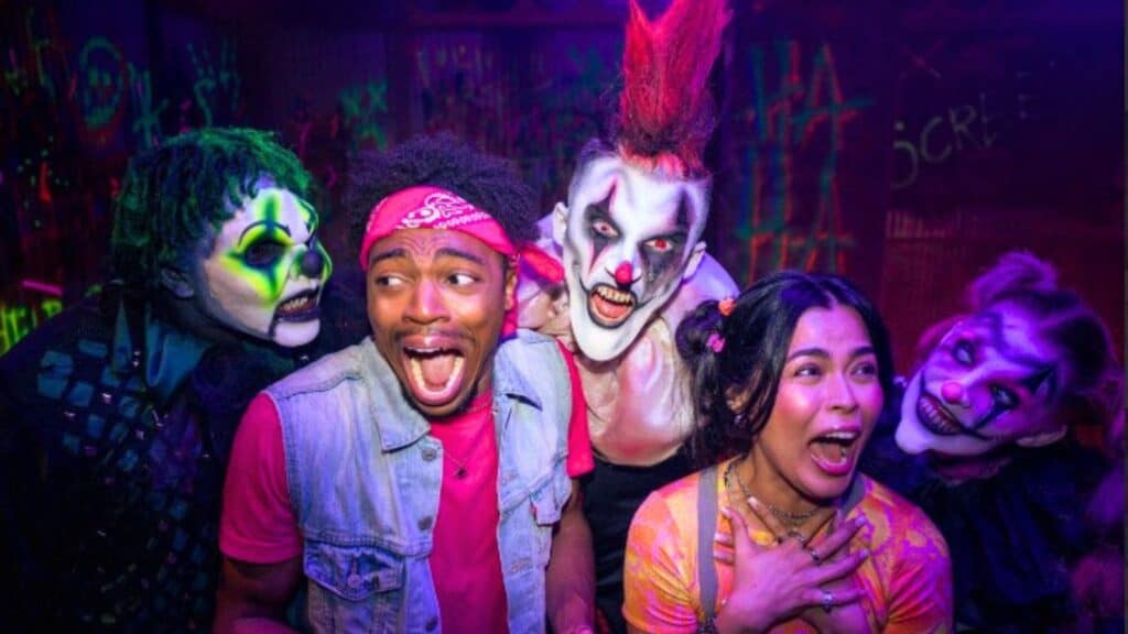 A man and woman look scared as they are surrounded by three people in creepy clown makeup under colorful lighting at the Six Flags 2026 Halloween event.