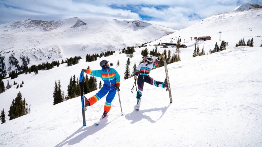 Two skiers in colorful outfits pose on a snowy mountain slope, each balancing on one ski with poles in hand. Snow-covered peaks and evergreen trees are visible in the background.
