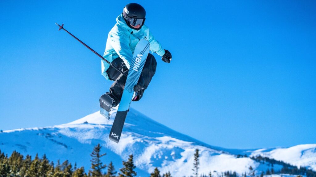 A skier wearing a blue jacket and helmet performs a mid-air trick with crossed skis above a snowy mountain landscape on a clear day.