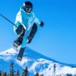 A skier wearing a blue jacket and helmet performs a mid-air trick with crossed skis above a snowy mountain landscape on a clear day.